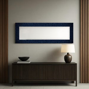 Puede incluir: Una pieza de arte mural rectangular con un marco azul marino estampado y un interior blanco. Debajo, un mueble de madera marrón oscuro con ranuras verticales contiene un cuenco negro, libros y una lámpara con una pantalla beige.