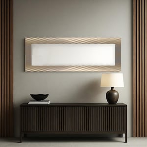 Op de afbeelding: Een rechthoekige spiegel met een getextureerde, goudkleurige lijst hangt boven een donkerbruin houten dressoir. Een lamp met een crèmekleurige kap staat op het dressoir, naast een zwarte kom en boeken. De muur is neutraal grijs.