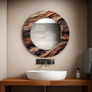 Puede incluir: Un espejo redondo con marco de madera natural, que muestra un patrón de vetas en tonos marrones y negros. Está montado sobre un lavabo ovalado blanco. El lavabo está sobre una superficie de madera, con un grifo negro y un dispensador de jabón.