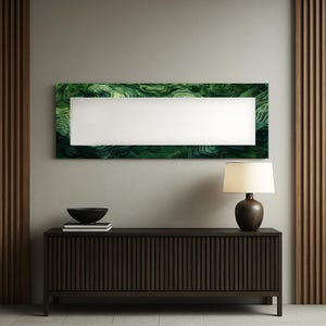 Op de afbeelding: Een lang, rechthoekig kunstwerk voor aan de muur met een wit midden en een groen, gelaagd, abstract ontwerp rond de randen. Daaronder een donkerbruin houten kast met verticale lijnen, een zwarte kom en een lamp met een beige kap.