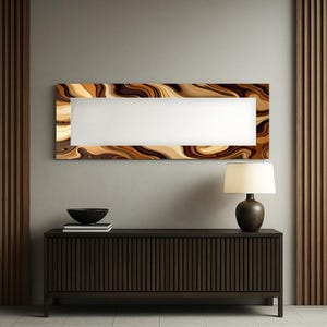 Op de afbeelding: Een rechthoekige wandspiegel met een wit midden en een decoratieve lijst met wervelende patronen in tinten bruin, beige en goud. De spiegel is gemonteerd op een grijze muur boven een donkerbruin dressoir. Een lamp en een zwarte kom zijn ook zichtbaar.