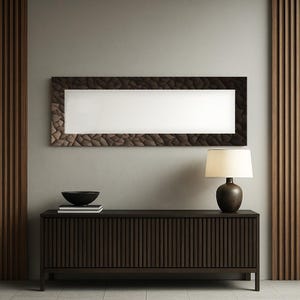 Puede incluir: Un espejo de pared rectangular con un marco marrón oscuro texturizado cuelga sobre un mueble de madera marrón oscuro. Un cuenco negro y libros están sobre el mueble. Una lámpara con una pantalla color crema y una base marrón oscuro está sobre el mueble.