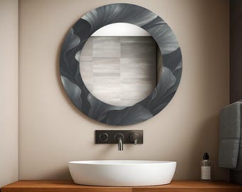 May include: A round mirror with a dark gray floral pattern frame hangs above a white oval sink. The sink sits on a wooden vanity with a black faucet and a small bottle. The mirror reflects a light-colored tiled wall.