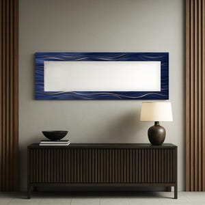 May include: A rectangular mirror with a navy blue frame featuring gold wave accents. The mirror hangs above a dark brown cabinet with a lamp and a black bowl. The wall is a neutral gray color.