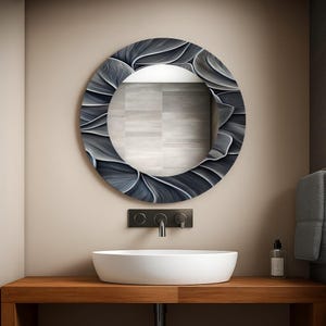 May include: A round, decorative mirror with a floral-like design in shades of gray and white. The mirror is mounted above a white, oval-shaped sink on a wooden vanity. A black faucet and wall-mounted controls are visible.