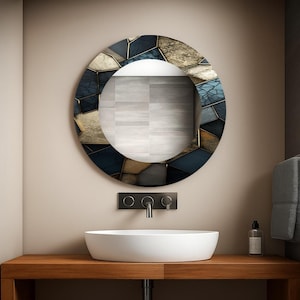 Puede incluir: Un espejo redondo decorativo con un patrón geométrico en tonos azules, dorados y grises. El espejo está montado sobre un lavabo ovalado blanco en una encimera de madera. También se ven un grifo negro y accesorios de pared.