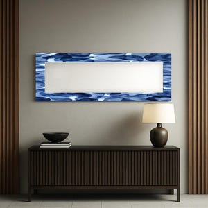 Puede incluir: Un espejo de pared rectangular con un marco de patrón de acuarela azul. El espejo está montado sobre un mueble de madera marrón oscuro. Un cuenco negro y una lámpara con base marrón y pantalla beige están sobre el mueble.