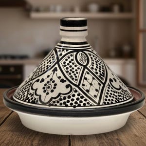 Peut inclure: Un tajine en céramique blanc et noir avec un couvercle conique. Le couvercle présente un motif géométrique noir et blanc détaillé. Le pot repose sur une base blanche avec un bord noir, idéal pour la cuisine marocaine.