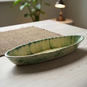 May include: A long, oval-shaped ceramic serving dish. The dish has a light yellow base with green streaks and a glossy finish. It sits on a light-colored wooden surface, suggesting a decorative or functional kitchen item.