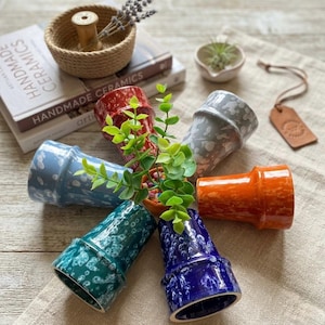 Peut inclure: Six pots de fleurs en céramique de différentes couleurs, dont le bleu, le rouge, l'orange et le vert, disposés en cercle. Chaque pot présente un glaçage moucheté unique. L'image comprend également un petit panier tressé, un livre et une étiquette en cuir.
