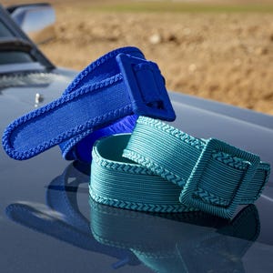 Puede incluir: Dos cinturones tejidos en tonos azules y turquesas. Los cinturones tienen una hebilla rectangular y un borde trenzado. Los cinturones se muestran sobre una superficie reflectante, con un fondo borroso de un coche y un campo.