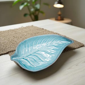 Peut inclure: Un plat de service en céramique turquoise en forme de feuille. Le plat présente une finition brillante et des nervures détaillées, ressemblant à une vraie feuille. Il repose sur un set de table tissé, suggérant un usage décoratif ou fonctionnel.
