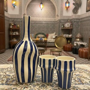 Peut inclure: Un vase blanc et deux tasses assorties, décorés de rayures bleues verticales. Le vase a un col fin et un corps arrondi. Les tasses sont cylindriques avec un dessus évasé. L'ensemble est posé sur une nappe à motifs.