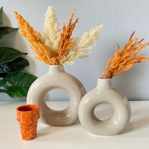 May include: Two beige ceramic vases with a circular opening, one large and one small, each holding dried orange and cream-colored pampas grass. A small orange ceramic pot is in front of the vases.