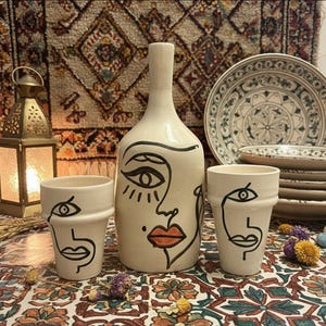 Peut inclure: Une bouteille en céramique crème et deux tasses assorties, chacune ornée d'un dessin de visage en noir. La bouteille a un long col et les tasses sont cylindriques. Des assiettes décoratives et une lanterne sont en arrière-plan.