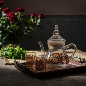 Puede incluir: Una tetera de cristal transparente y dos vasos a juego sobre una bandeja de madera. La tetera está decorada con motivos florales rojos, verdes y dorados. Un ramo de rosas rojas y menta fresca se encuentra en el fondo.