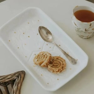 Peut inclure: Un plateau de service rectangulaire blanc avec une cuillère en argent et deux biscuits en spirale. Le plateau présente un motif en relief subtil et est parsemé de points dorés. Une tasse de thé et un panier en osier sont également visibles.