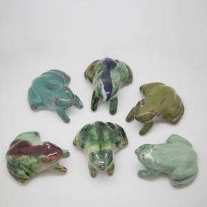 Könnte beinhalten: Sechs Keramikfroschfiguren in verschiedenen Grün-, Blau- und Marontönen. Jeder Frosch hat ein einzigartiges Glasurmuster und befindet sich in einer entspannten Pose. Ideal für die Inneneinrichtung.