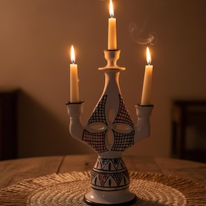 Peut inclure: Un chandelier en céramique blanche avec trois bougies allumées. Le chandelier présente un motif décoratif noir et rouge. Les bougies sont de couleur ivoire et les flammes sont vives. Le chandelier est posé sur un dessous de plat rond et tissé.