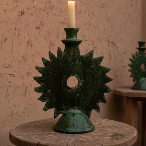 Peut inclure: Un bougeoir en céramique verte avec un motif en forme d'éclat d'étoile et une bougie blanche allumée. Le bougeoir a une ouverture circulaire au centre et repose sur une petite table en bois. Un second bougeoir est visible en arrière-plan.