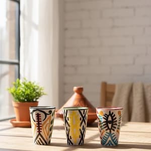 Peut inclure: Trois verres à boire en céramique avec des motifs géométriques colorés. Les verres ont une forme cylindrique et sont décorés de motifs noirs, jaunes, orange et bleus. Les verres sont sur une table en bois, avec un pot en terre cuite et un tajine en arrière-plan.