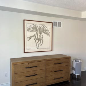 May include: A wooden dresser with six drawers and black handles. Above it hangs a framed artwork depicting a winged horse. The room has white walls, a dark wood floor, and a small air purifier.