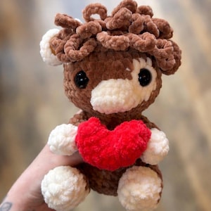 May include: A crochet cow plush toy, holding a red heart. The toy is made of brown and white yarn, with black eyes. The heart is a bright red colour. A charming and cuddly gift for Valentine's Day or any occasion.