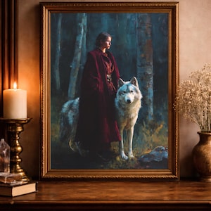 May include: A framed artwork depicts a woman in a red cloak standing with a large wolf in a forest setting. The painting has a gold frame and is displayed on a wooden table. A lit candle and a vase of dried flowers are also visible.