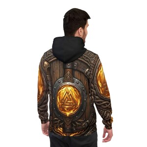 Peut inclure: Un sweat à capuche avec un motif détaillé d'un bouclier en bois avec un emblème central et des ornements en métal. Le design incorpore des tons chauds de marron, d'or et de bronze, avec une capuche noire.
