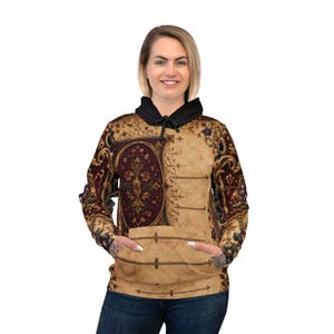 May include: A beige hoodie with a black hood and a large front pocket. The hoodie features a detailed, ornate design in burgundy, gold, and brown, resembling a tapestry or antique book cover. The person is wearing blue jeans.