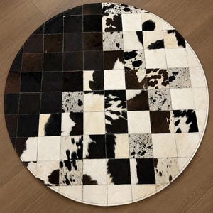 May include: A round cowhide rug with a patchwork design. The rug features a checkerboard pattern of black, white, and brown squares. The cowhide has a natural texture and a stitched border. The rug is on a wooden floor.