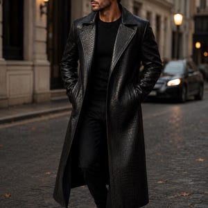 Op de afbeelding: Een lange, zwarte leren jas met een getextureerde afwerking, die op krokodillenleer lijkt. De jas heeft een klassieke reverskraag en wordt gedragen met zwarte kleding. De persoon draagt een zonnebril en zwarte schoenen en loopt op een geplaveide straat.