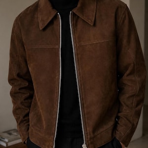 Puede incluir: Chaqueta de ante marrón con cuello y cremallera plateada. La chaqueta se lleva sobre un cuello alto negro. La chaqueta tiene un diseño clásico y un corte entallado, adecuado para diversas ocasiones.