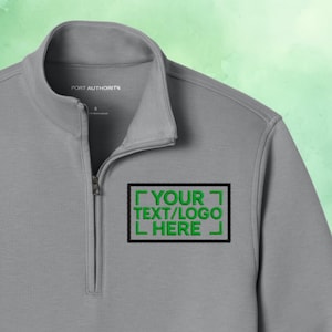 Puede incluir: Un jersey gris con cremallera de cuarto de cierre con el texto "YOUR TEXT/LOGO HERE" bordado en verde dentro de un rectángulo negro. La camisa tiene un cuello alzado y una cremallera que se extiende por el pecho. La marca "PORT AUTHORITY" está impresa en el cuello.
