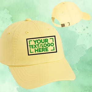Puede incluir: Gorra de béisbol amarillo claro con visera curva. La gorra presenta un diseño rectangular verde y negro con el texto "YOUR TEXT/LOGO HERE". Tiene un cierre metálico para ajustar.