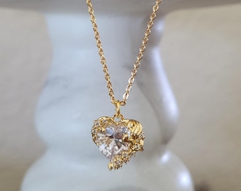 May include: A gold-colored necklace with a heart-shaped pendant. The pendant features a large, clear gemstone at its center, surrounded by smaller, decorative elements. The necklace hangs against a white marble pedestal, with dried flowers in the background.