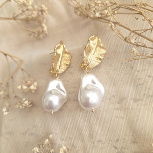 May include: A pair of gold-colored leaf-shaped earrings with large, irregular white pearl drops. The earrings are set against a soft, neutral-toned background with delicate dried flowers.