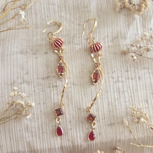 May include: A pair of gold-toned earrings with a cascading design. Each earring features a red and gold striped bead, a teardrop-shaped red gem, and a square-cut red gem. The earrings are set against a soft, textured background with delicate floral accents.
