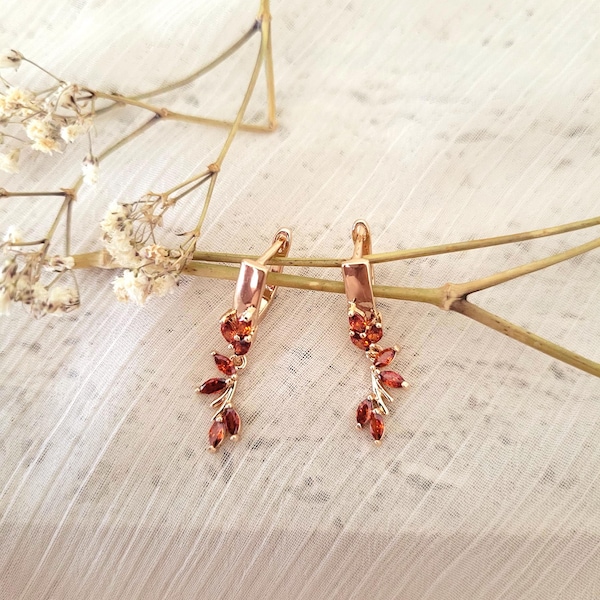 May include: A pair of gold-colored earrings with a floral design. The earrings feature a rectangular top with a series of small, reddish-orange gemstones arranged to resemble leaves and flowers. The earrings are displayed on a white surface.