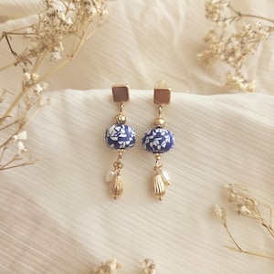 May include: A pair of dangle earrings featuring blue and white patterned ceramic beads. The earrings have gold-colored accents, including square studs, small beads, and teardrop-shaped dangles with small white pearls.