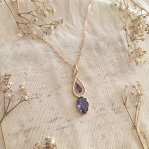 May include: A rose gold necklace with a teardrop pendant featuring two blue gemstones. The necklace is displayed on a white, textured fabric, with dried flowers in the background. The pendant has a small gemstone above a larger one.