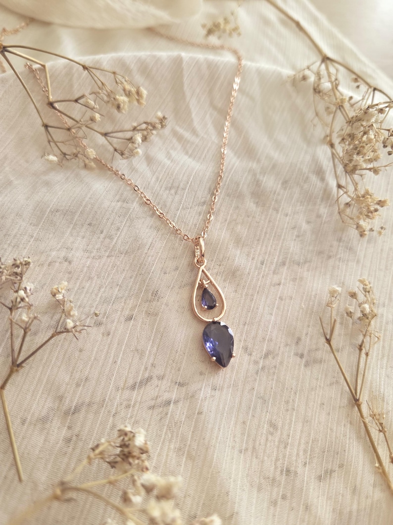 May include: A rose gold necklace with a teardrop pendant featuring two blue gemstones. The necklace is displayed on a white, textured fabric, with dried flowers in the background. The pendant has a small gemstone above a larger one.