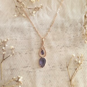 May include: A rose gold necklace with a teardrop pendant featuring a small, round, and a large, teardrop-shaped, dark purple gemstone. The necklace is displayed on a white, textured surface, with dried flowers in the background.