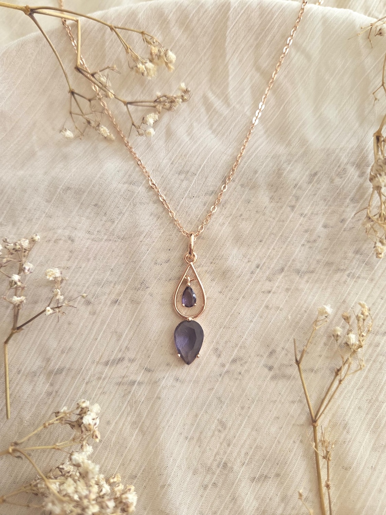May include: A rose gold necklace with a teardrop pendant featuring a small, round, and a large, teardrop-shaped, dark purple gemstone. The necklace is displayed on a white, textured surface, with dried flowers in the background.