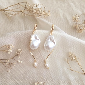 May include: A pair of gold-tone hoop earrings featuring a dangling, irregular white baroque pearl and a slender, spiraling gold-tone wire that terminates in a small, round white pearl at the bottom of each earring.