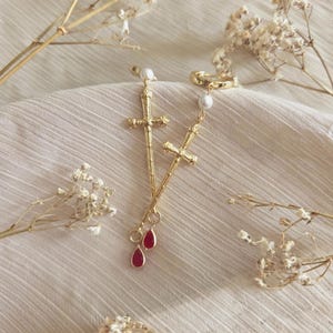 May include: Gold-colored sword earrings with a cross design and red teardrop accents. Each earring features a pearl at the top. The earrings are displayed on a cream-colored fabric with dried flowers.