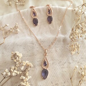 May include: A rose gold jewelry set featuring a necklace and matching earrings. The necklace has a teardrop pendant with a dark purple gemstone. The earrings also have a teardrop design with a similar gemstone. The set is displayed on a white fabric.
