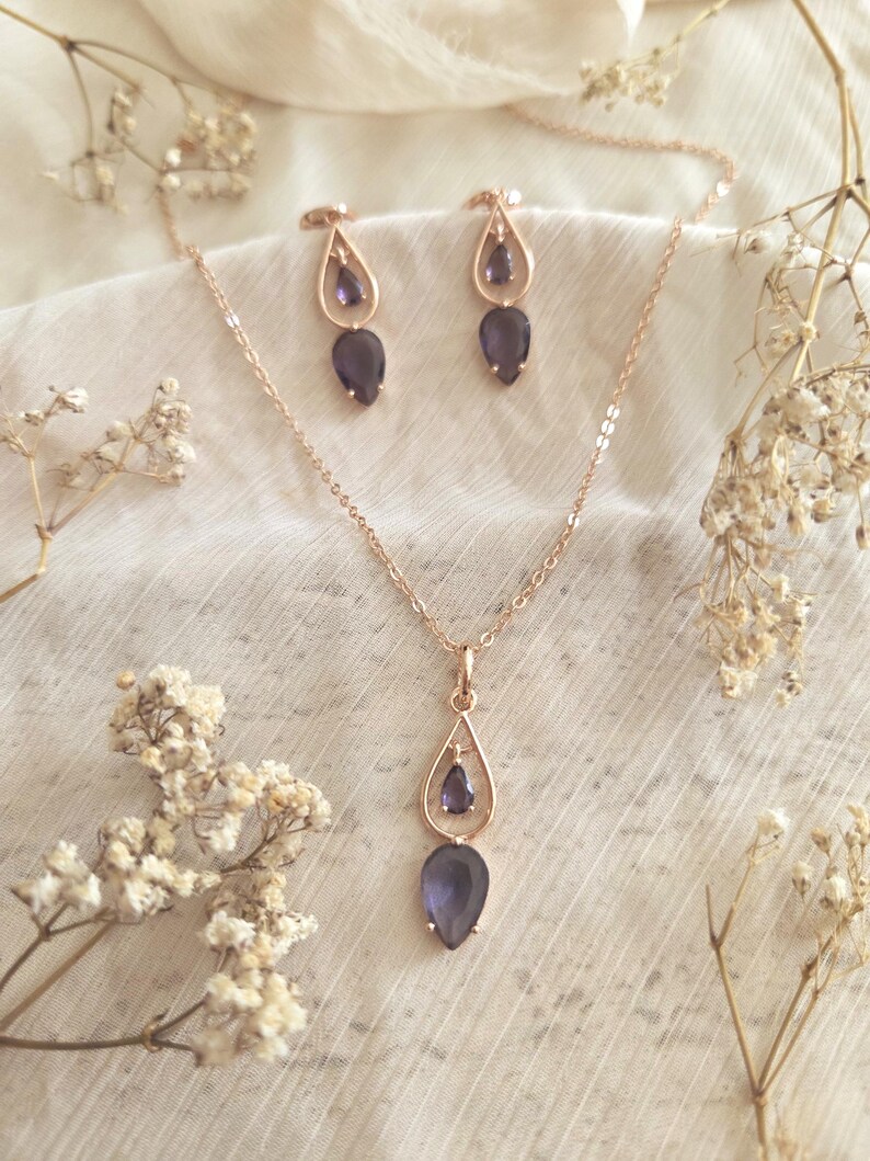 May include: A rose gold jewelry set featuring a necklace and matching earrings. The necklace has a teardrop pendant with a dark purple gemstone. The earrings also have a teardrop design with a similar gemstone. The set is displayed on a white fabric.