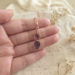 May include: A gold-colored necklace with a teardrop-shaped purple gemstone pendant. The pendant is suspended from a delicate chain and is held in a hand against a soft, neutral background. The necklace has a simple, elegant design.