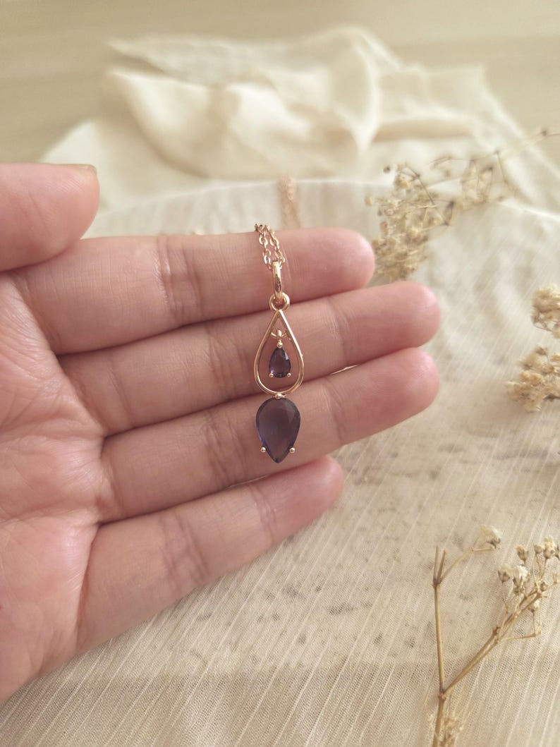 May include: A gold-colored necklace with a teardrop-shaped purple gemstone pendant. The pendant is suspended from a delicate chain and is held in a hand against a soft, neutral background. The necklace has a simple, elegant design.